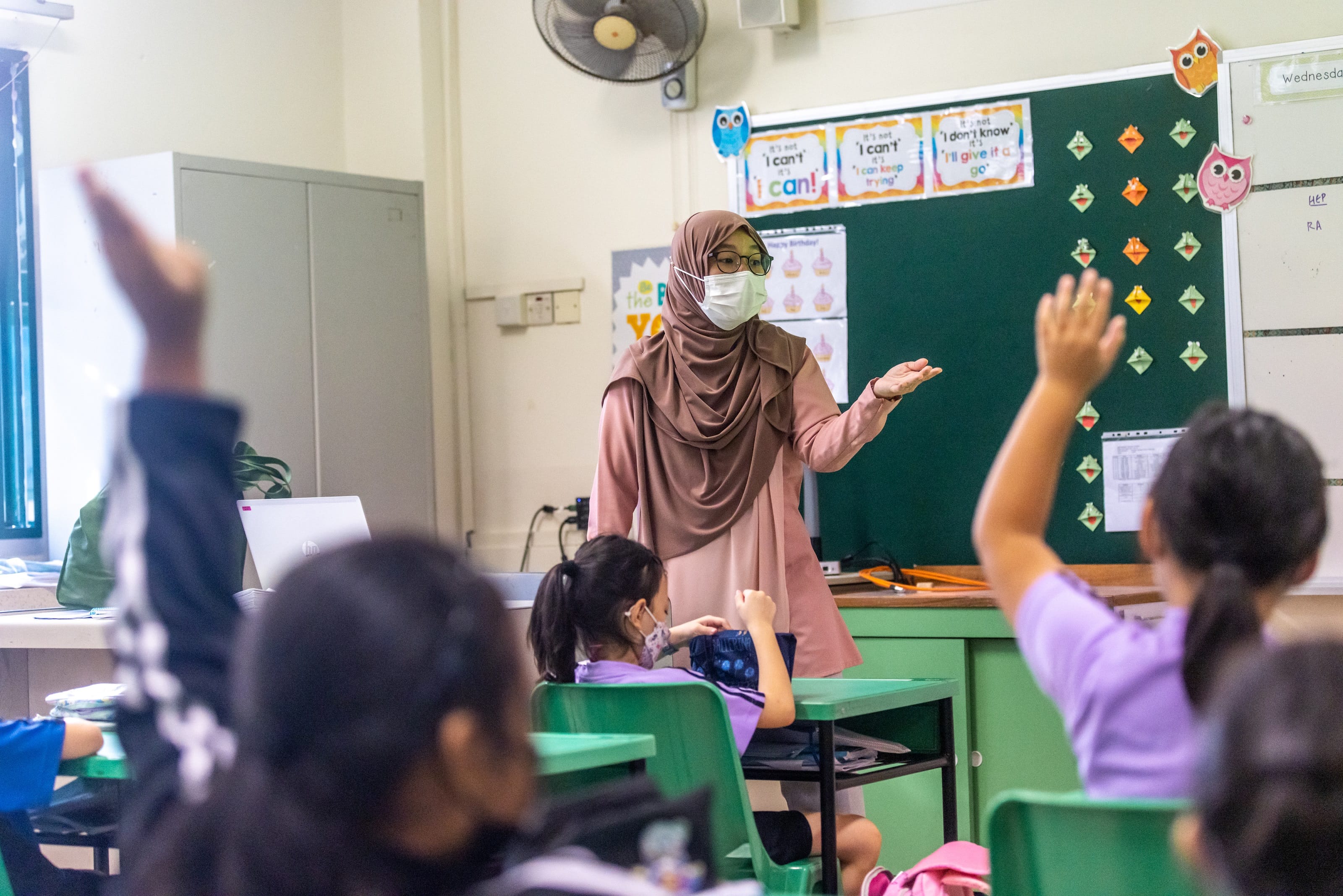 A masked teacher wearing a hijab stands at a green chalkboard as young students raise their hands in a decorated classroom.